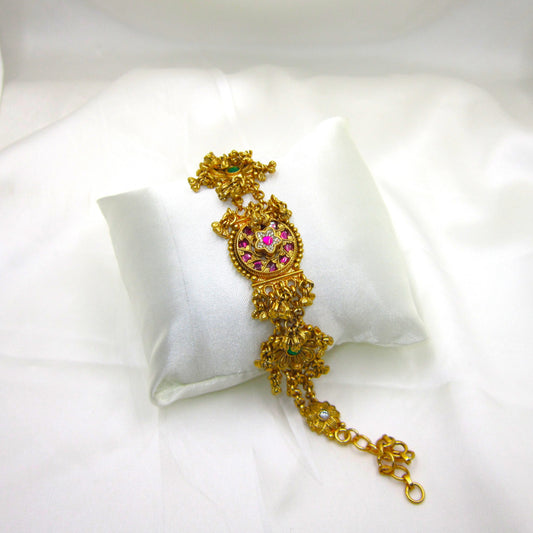 Alankrita Royal Ruby Pink Bracelet by Enava featuring antique gold finish, ruby pink stones, emerald green floral accents, American Diamond detailing, intricate heritage craftsmanship, hanging traditional charms, adjustable chain closure, and a premium ethnic statement bracelet for weddings, festive occasions, and bridal wear.