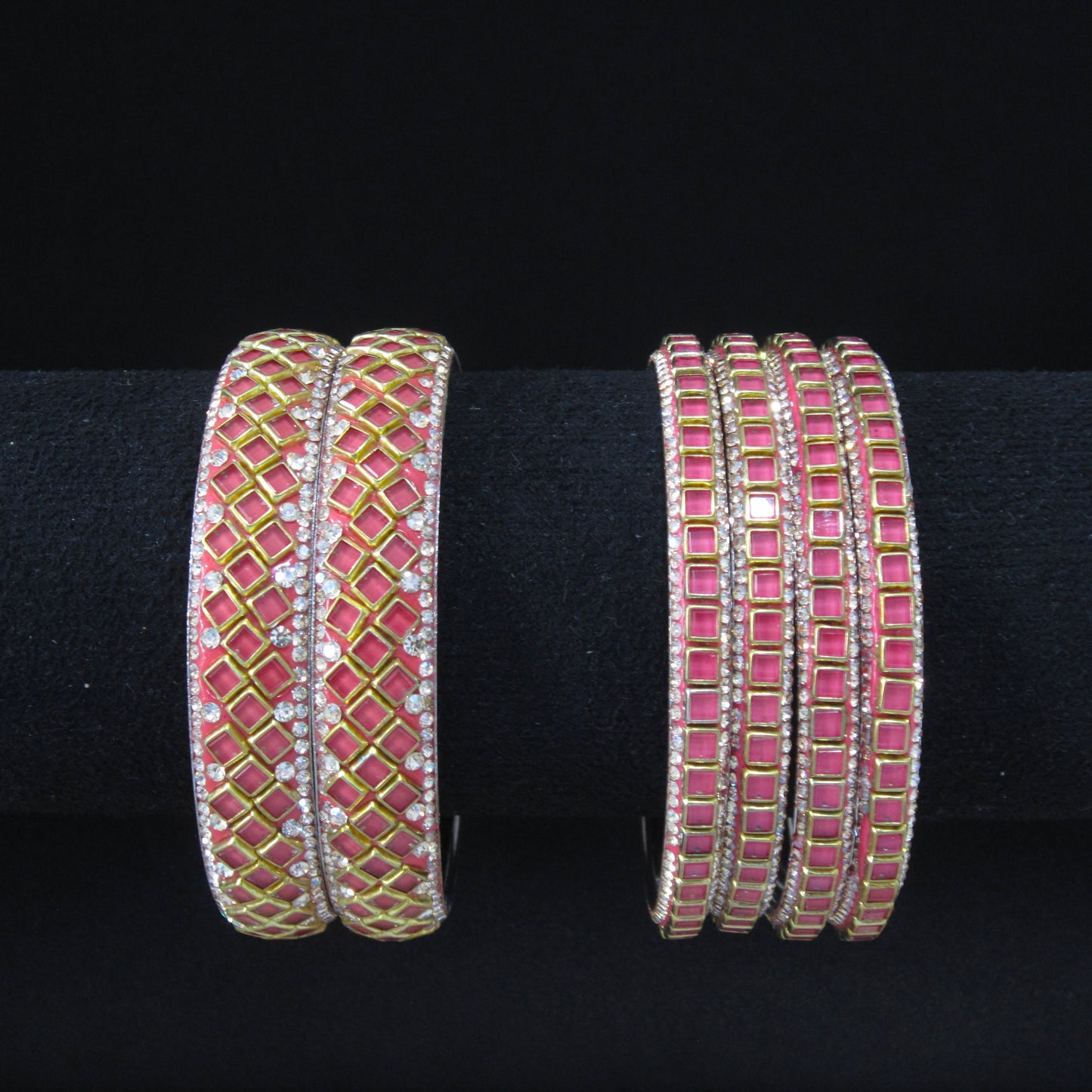 Two pairs of gold-plated bangles decorated with pink and clear rhinestones, displayed on a black velvet ring stand against a black background.