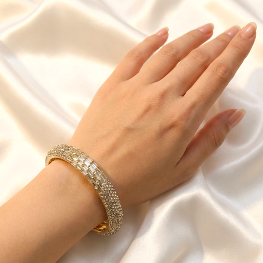 A close-up of a hand wearing a wide, gold-toned bangle bracelet covered in clear rhinestones, resting on draped white satin fabric.