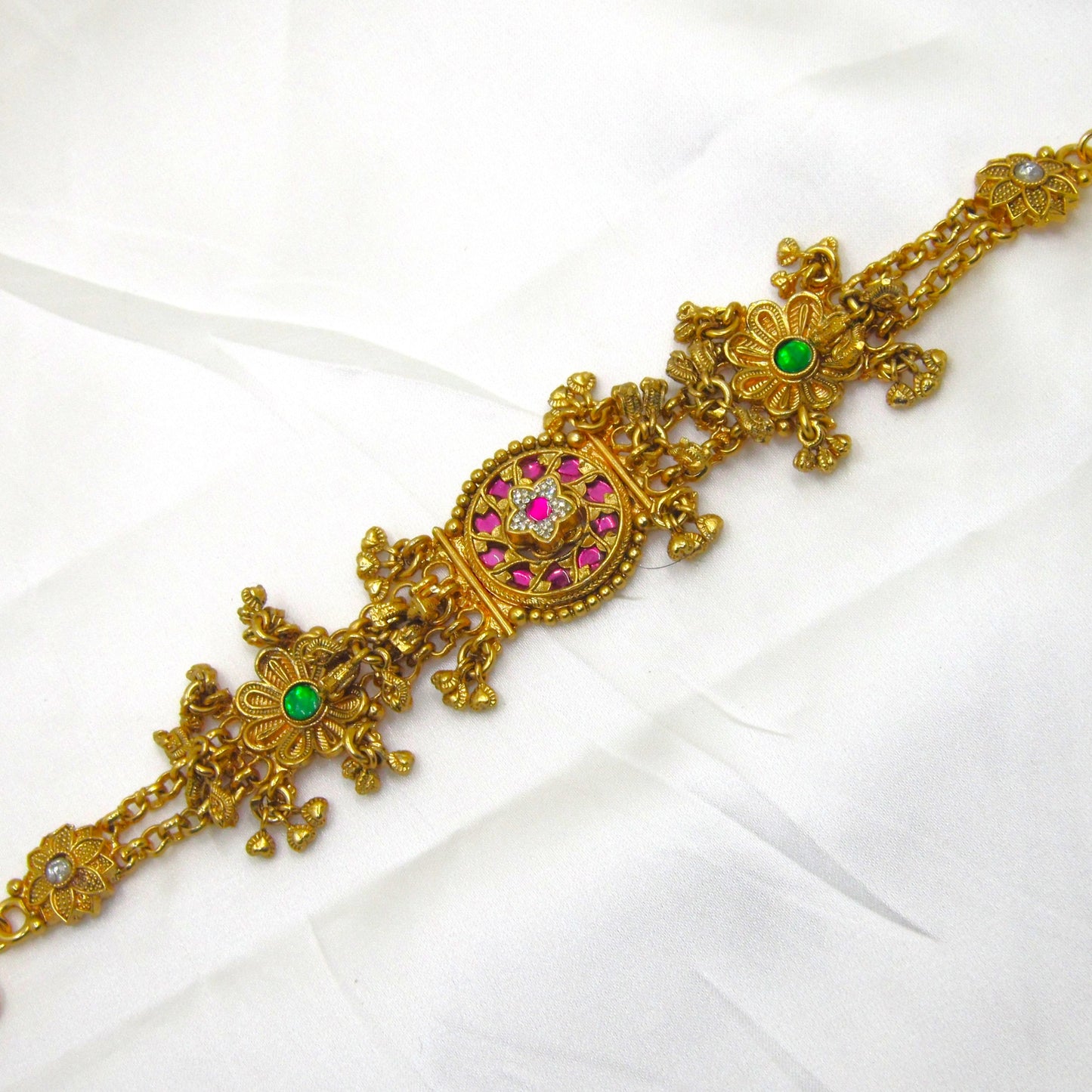 Alankrita Royal Ruby Pink Bracelet by Enava featuring antique gold finish, ruby pink stones, emerald green floral accents, American Diamond detailing, intricate heritage craftsmanship, hanging traditional charms, adjustable chain closure, and a premium ethnic statement bracelet for weddings, festive occasions, and bridal wear.