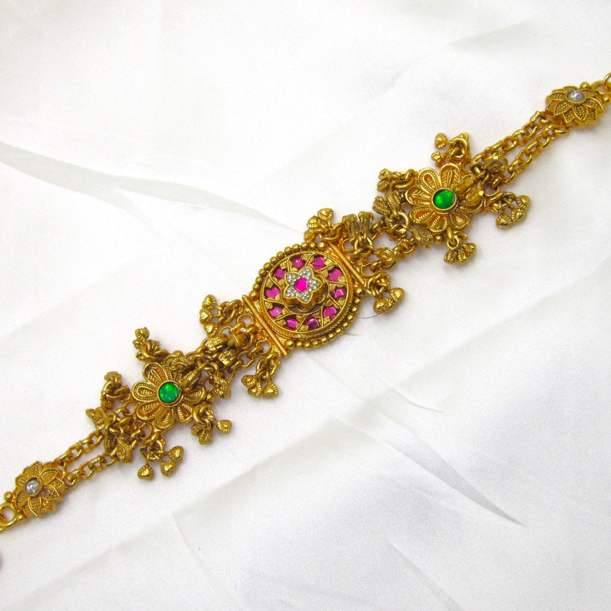 Alankrita Royal Ruby Pink Bracelet by Enava featuring antique gold finish, ruby pink stones, emerald green floral accents, American Diamond detailing, intricate heritage craftsmanship, hanging traditional charms, adjustable chain closure, and a premium ethnic statement bracelet for weddings, festive occasions, and bridal wear.