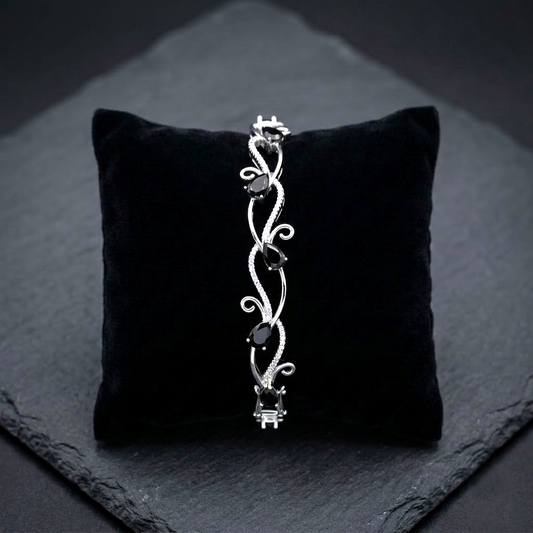 A silver-toned bangle bracelet with three dark teardrop-shaped stones and intricate scrollwork, displayed on a black velvet cushion over a dark slate surface.