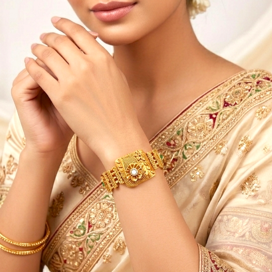 A close-up of a woman wearing an ornate gold bracelet on her wrist, paired with a matching gold bracelet. She is wearing a cream-colored embroidered saree, set against a soft, light background.