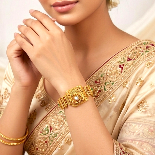 A close-up of a woman wearing an ornate gold bracelet on her wrist, paired with a matching gold bracelet. She is wearing a cream-colored embroidered saree, set against a soft, light background.