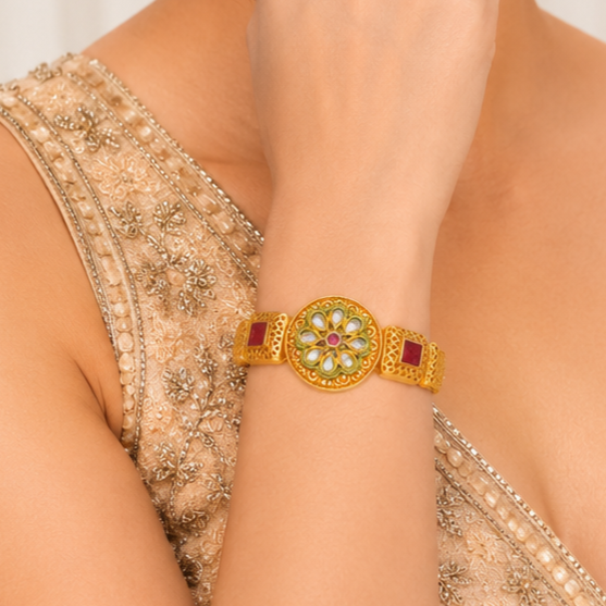 A close-up of a woman's wrist wearing a traditional Indian gold bracelet (kara) decorated with a central floral motif featuring light green stones, a small pink center, and red square side stones.