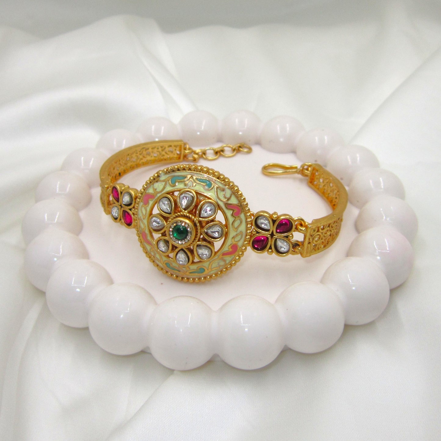 Kalashini Enamel Ethnic Bracelet by Enava featuring a circular Kundan centerpiece with hand-painted ivory Meenakari enamel, emerald green center stone, ruby pink floral accents, antique gold finish, adjustable closure, and a premium ethnic statement bracelet for weddings and festive occasions.