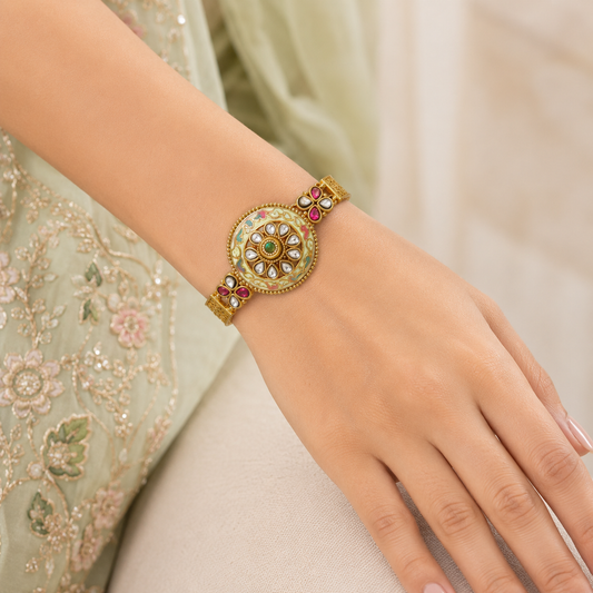 Kalashini Enamel Ethnic Bracelet by Enava featuring a circular Kundan centerpiece with hand-painted ivory Meenakari enamel, emerald green center stone, ruby pink floral accents, antique gold finish, adjustable closure, and a premium ethnic statement bracelet for weddings and festive occasions.