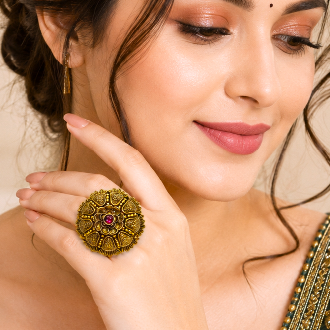 A close-up lifestyle shot of a woman wearing a large, ornate gold-toned ring with a central pink gemstone on her finger. She is also wearing a matching gold earring.