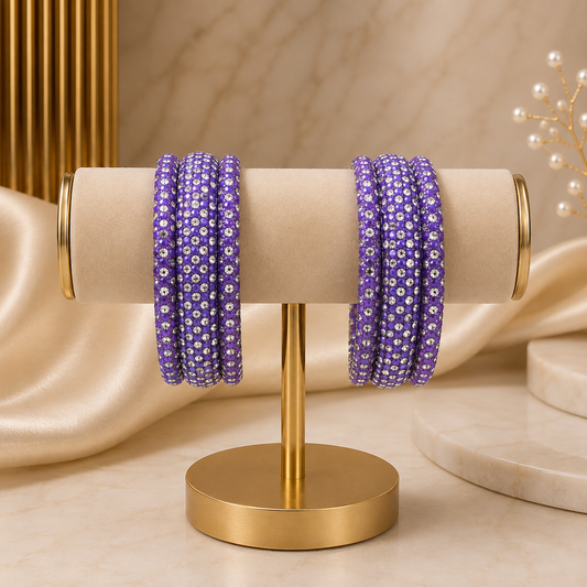 Purple beaded bangles with white floral patterns displayed on a gold stand against a beige marble and satin background.