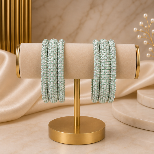 Two sets of beaded bangles in a pale mint green color, displayed on a gold stand with a beige velvet sleeve. The background features draped satin fabric, marble surfaces, and gold decorative columns.