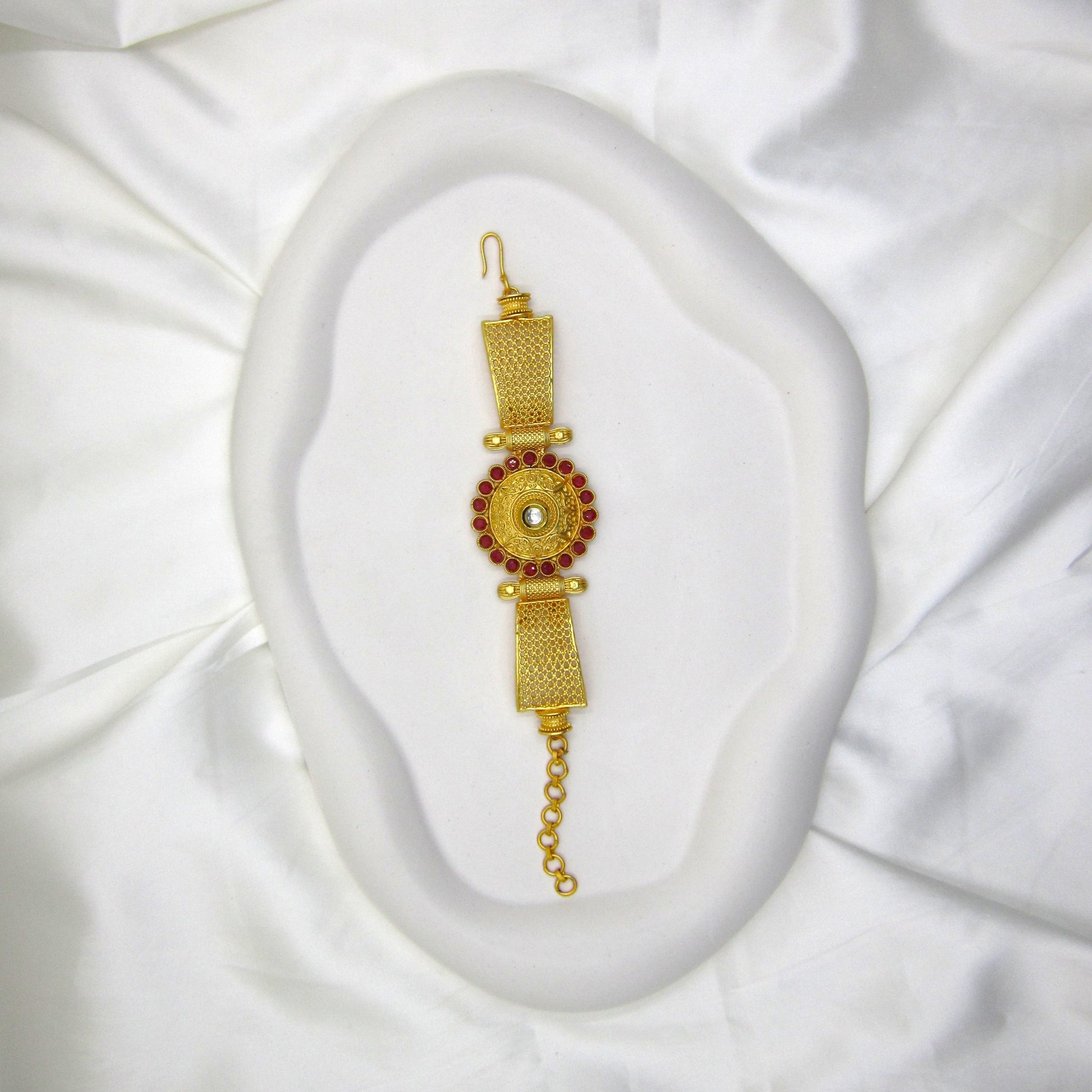 Mrinalini Ruby Golden Bracelet by Enava featuring an engraved antique gold circular centerpiece with a mirror stone, ruby red crystal border, textured mesh band, adjustable chain closure, premium ethnic statement bracelet for weddings and festive wear.