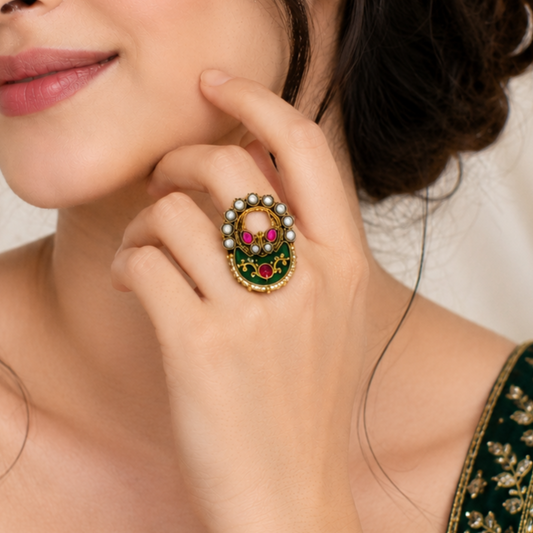 A close-up lifestyle shot of a woman wearing a highly detailed gold ring with green enamel, red stones, and white pearls, set against her neck and hand.