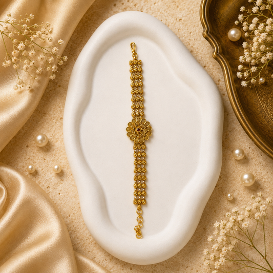 A gold-toned traditional Indian bracelet (bangle) with a detailed central floral motif and beaded chain, displayed on a white wavy jewelry stand. The stand is set against a beige textured background decorated with gold satin fabric, white pearls, and dried baby's breath flowers.