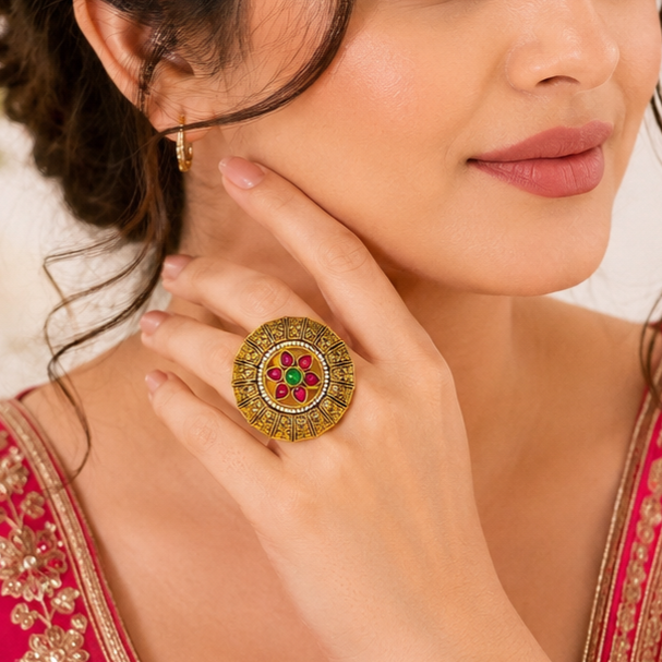A close-up lifestyle shot of a woman wearing an ornate gold ring with red and green stone accents on her finger, along with a matching gold earring.