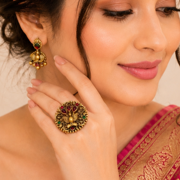A close-up lifestyle shot of a woman wearing an ornate gold ring and matching gold jhumka earrings with red and green stone accents.
