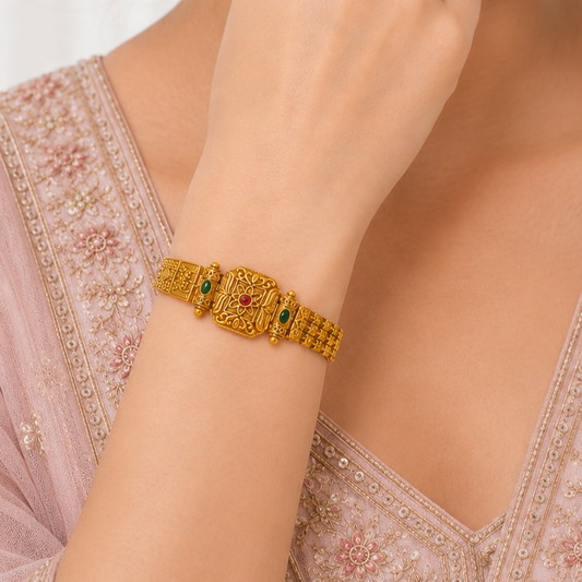 A close-up of a person's wrist wearing an ornate gold-toned traditional bracelet with small green and red stone accents, paired with a light pink embroidered top.