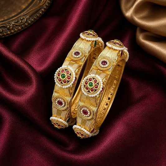 Two ornate gold bangles with red and green stone accents and white pearl-like borders, displayed on draped burgundy satin fabric.