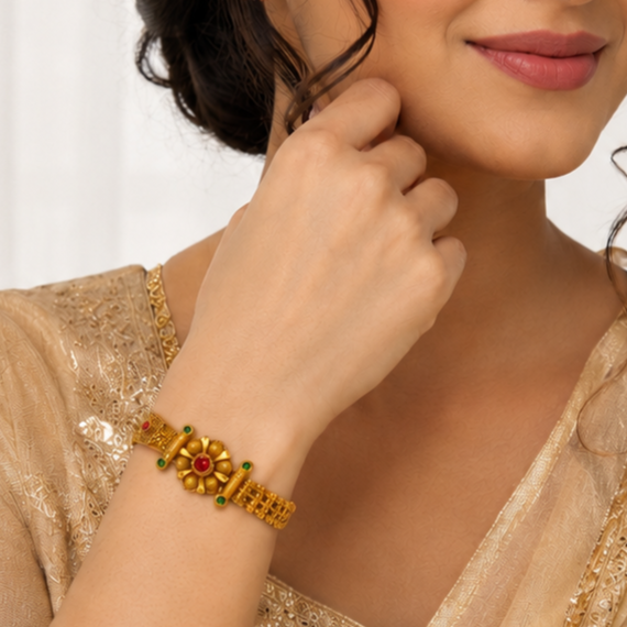 A close-up lifestyle shot of a woman wearing a gold-plated traditional Indian bracelet (kara) with a central floral motif featuring a red stone and green stones, paired with a beige embroidered top.