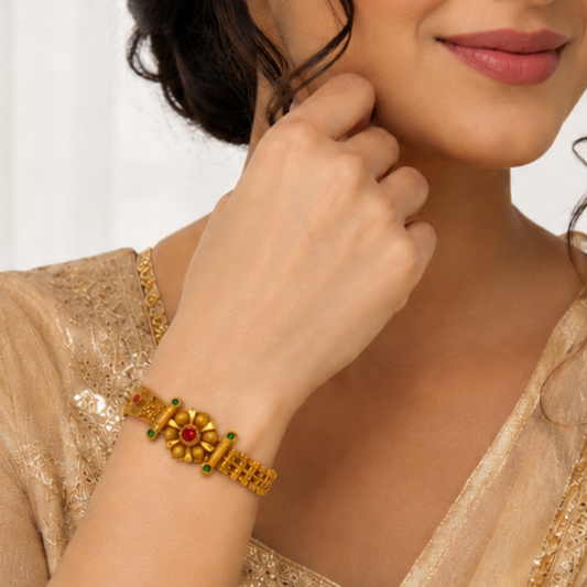 A close-up lifestyle shot of a woman wearing a gold-plated traditional Indian bracelet (kara) with a central floral motif featuring a red stone and green stones, paired with a beige embroidered top.