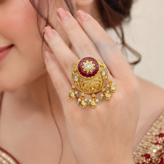 A close-up of a woman's hand wearing a large ornate gold ring with a deep red circular center, white floral detailing, and dangling gold charms with small green and white stones.