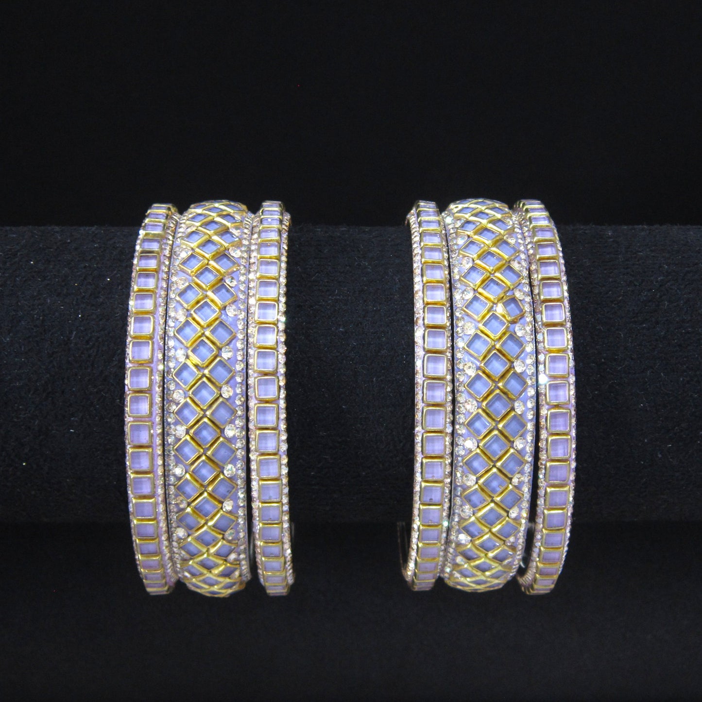 A pair of wide gold-plated bangles decorated with light purple iridescent stones and small clear rhinestone accents, displayed on a black ring stand against a black background.