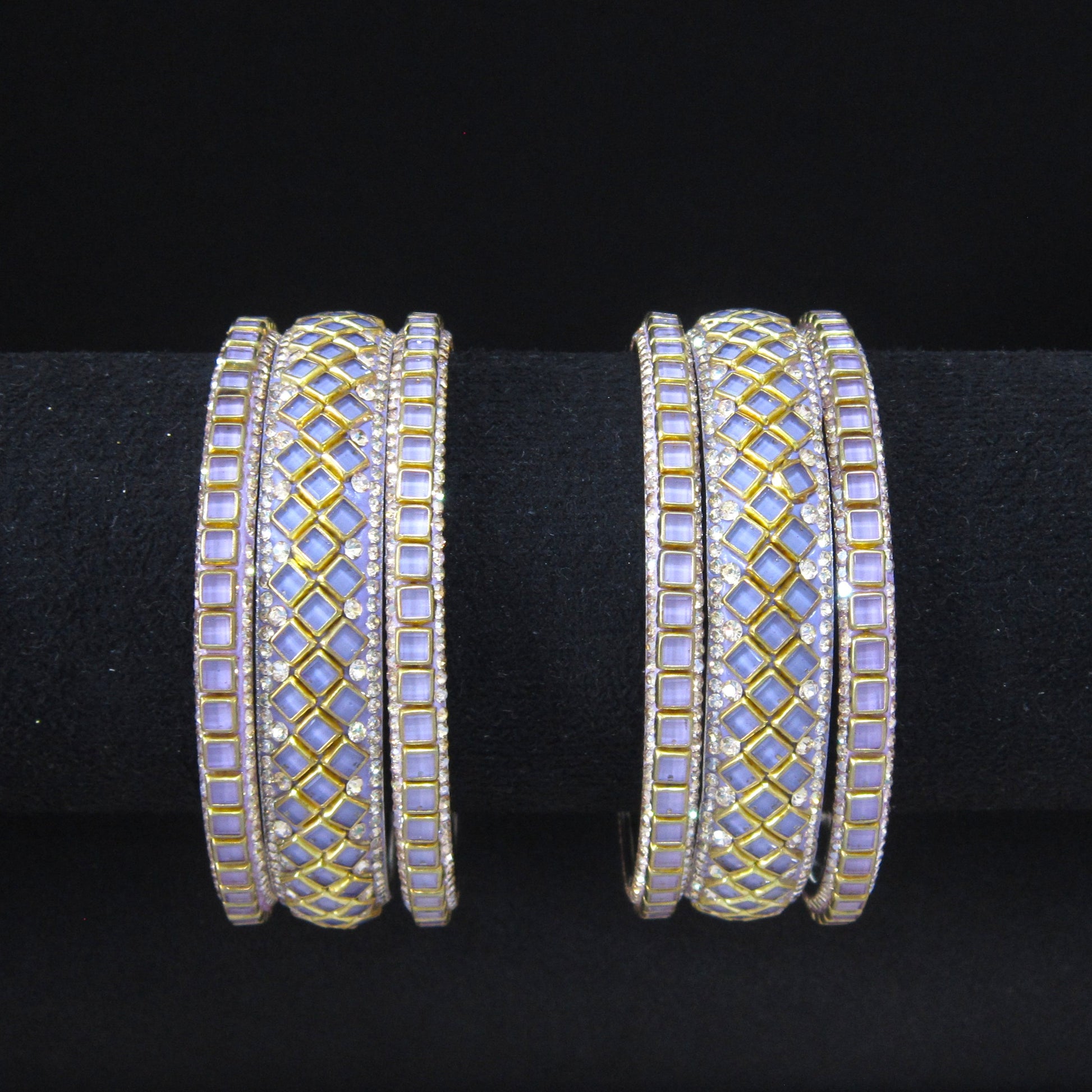 A pair of wide gold-plated bangles decorated with light purple iridescent stones and small clear rhinestone accents, displayed on a black ring stand against a black background.