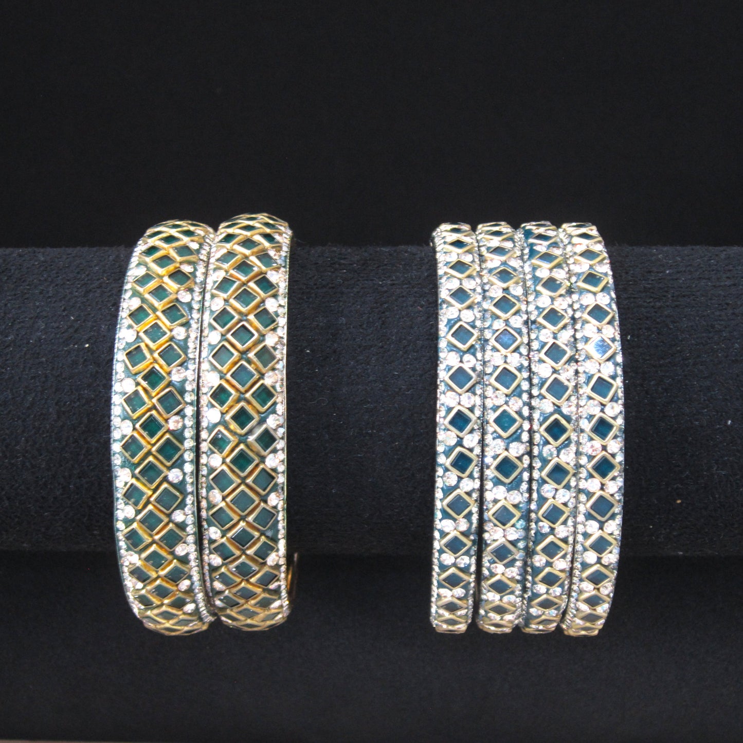 Two sets of multi-strand diamond-embellished bangles displayed on a black velvet jewelry stand against a black background. The left set features green stone accents with gold-toned metal borders, while the right set features blue-green stone accents with silver-toned metal borders.