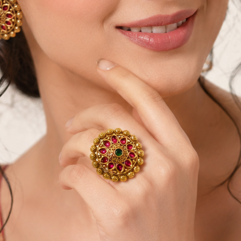 A close-up lifestyle shot of a woman wearing a large, ornate gold ring with red and green gemstones on her finger, along with matching gold earrings.